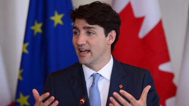 Un homme aux cheveux bruns est en train de parler en gesticulant des mains. Derrière lui, on peut voir le drapeau de l'Europe à côté de celui canadien.