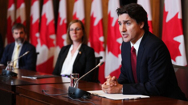 Justin Trudeau, avec deux ministres à sa droite, parle assis devant un micro, avec plusieurs drapeaux du Canada derrière lui.