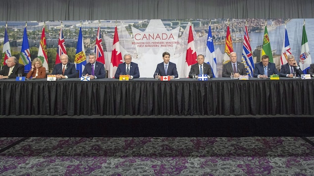 Plusieurs personnes sont assises à une même longue table devant les drapeaux des différentes provinces et territoires canadiens lors d'une conférence de presse.