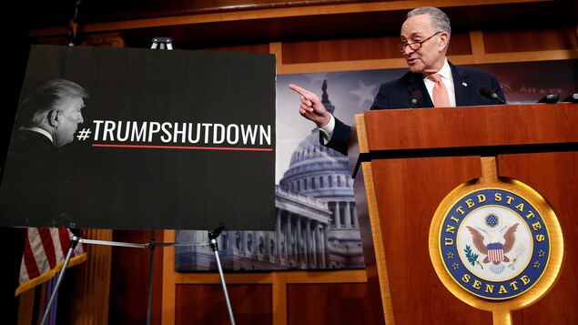 Chuck Shumer parle à un podium et pointe vers une pancarte sur laquelle on peut voir le visage de Donald Trump et le mot-clic #Trumpshutdown. Chuck Shumer parle à un podium et pointe vers une pancarte sur laquelle on peut voir le visage de Donald Trump et le mot-clic #Trumpshutdown.