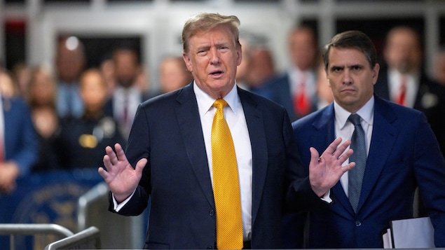 Donald Trump, qui porte une cravate jaune, s'adresse aux journalistes, les mains levées.