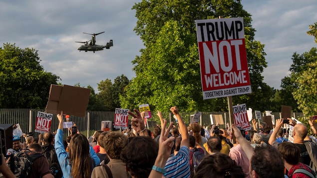 Des dizaines de personnes lèvent leur poing en direction d'un hélicoptère qui passe devant eux. Les manifestants brandissent aussi des pancartes disant « Trump n'est pas le bienvenu ». Des dizaines de personnes lèvent leur poing en direction d'un hélicoptère qui passe devant eux. Les manifestants brandissent aussi des pancartes disant « Trump n'est pas le bienvenu ».
