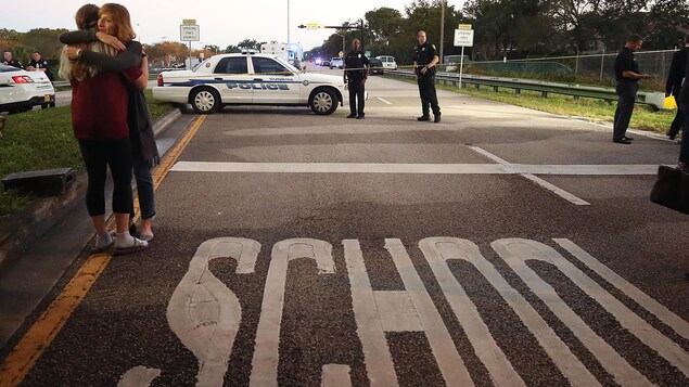 Deux femmes s'enlacent, près d'une voiture de police et de deux policiers. L'inscription « École » apparaît sur la chaussée. Deux femmes s'enlacent, près d'une voiture de police et de deux policiers. L'inscription « École » apparaît sur la chaussée.