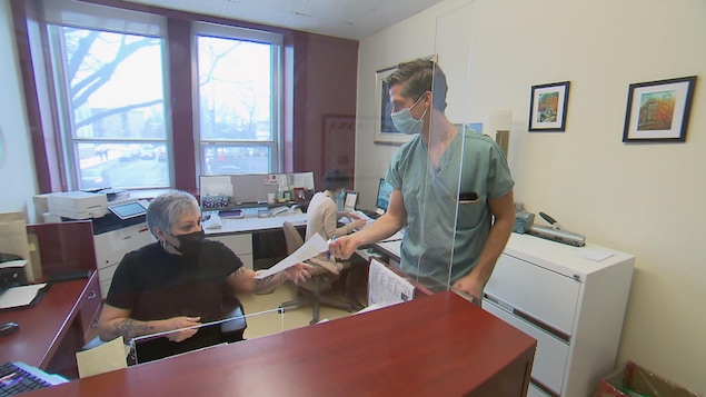 Un homme en blouse de médecin tend une feuille à une famme assise à un bureau.
