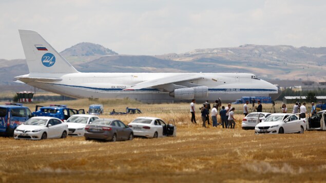 Un Antonov 124 de l'armée russe est visible derrière plusieurs véhicules. Des journalistes sont présents. 