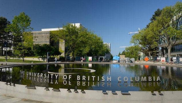 Des lettres (University of British Columbia) se reflètent dans l'eau d'une fontaine, avec des arbres et des bâtiments an arrière-plan.