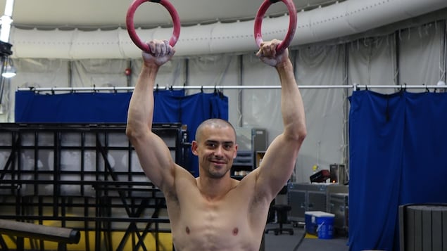 La photo montre un homme qui sourit, les mains relevées et tenant des anneaux de cirque.