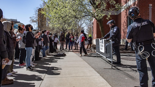 Des dizaines d'étudiants sont debout devant une barrière près de laquelle des policiers interdisent l'accès au site.