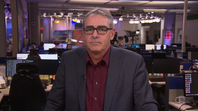 Un homme portant un veston et des lunettes rectangulaires regarde droit devant lui. Nous le voyons de face.