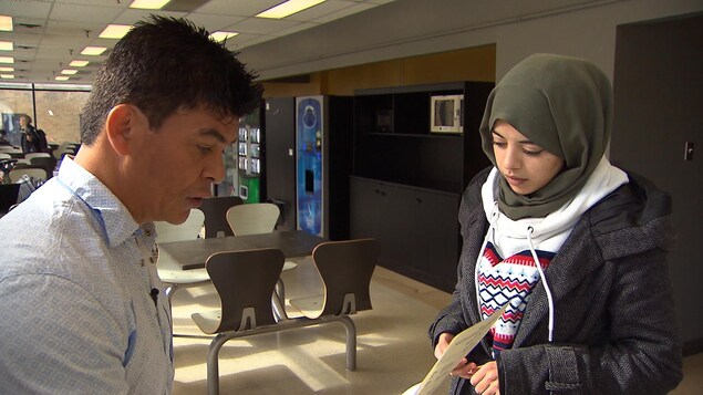 Un homme tend un formulaire à une jeune femme portant un hijab et un manteau dans une cafétéria de l'université.