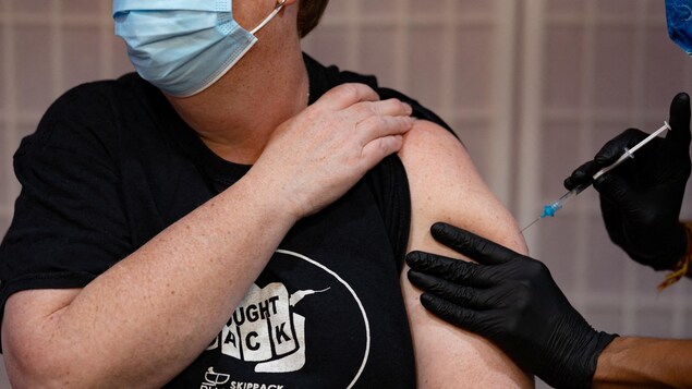 Une femme assise la tête tournée vers la droite relève la manche gauche de son t-shirt noir et reçoit un vaccin.