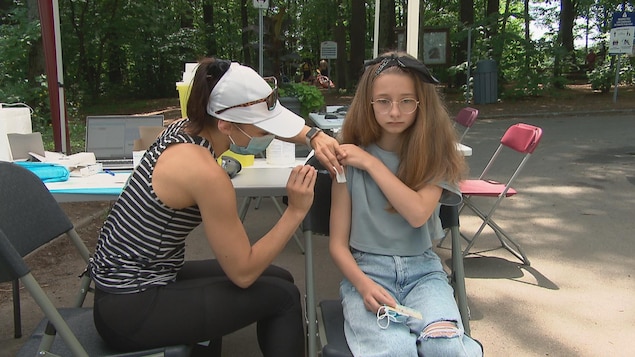 Une femme qui porte un masque chirurgical est en train d'injecter une dose de vaccin dans le bras d'une jeune fille. Toutes les deux sont assises, la scène se passe en extérieur.