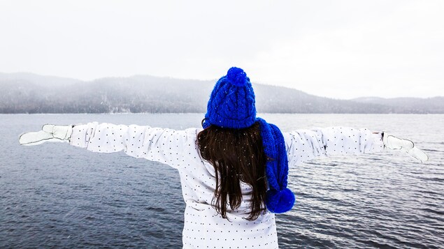Valérie Jessica Laporte de dos bras écartés face à une étendue d'eau, vêtue d'un manteau et de mitaines blanches, avec une tuque et un foulard bleus. Valérie Jessica Laporte de dos bras écartés face à une étendue d'eau, vêtue d'un manteau et de mitaines blanches, avec une tuque et un foulard bleus.