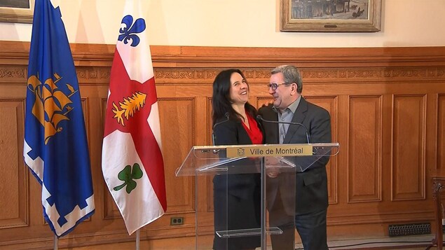 Les deux maires, debout derrière un lutrin, ricanent en se regardant l'un l'autre. Les drapeaux des Villes de Québec et Montréal sont visibles, à gauche de l'image.