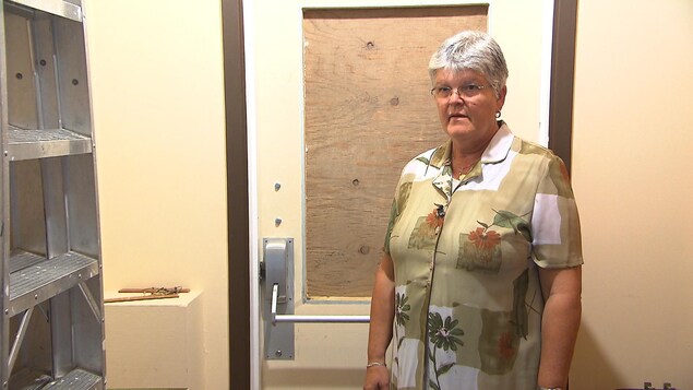 La directrice se trouve devant la porte dont la fenêtre a été brisée par les vandales et remplacée par un morceau de bois. La directrice se trouve devant la porte dont la fenêtre a été brisée par les vandales et remplacée par un morceau de bois.