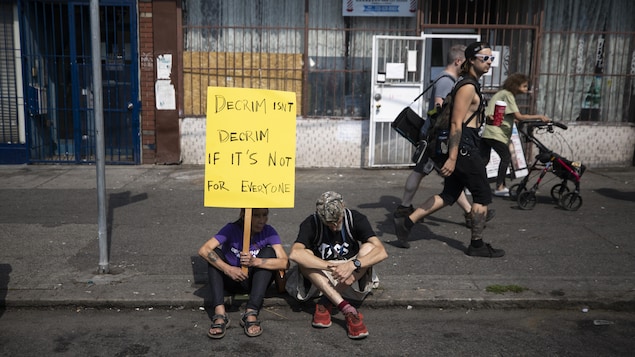 Deux personnes sont assises sur le trottoir, dont une femme qui tient une pancarte sur la décriminalisation.