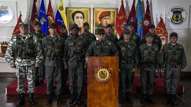 Plusieurs militaires en uniforme se tiennent bien droits de chaque côté d'un lutrin où un homme parle à des journalistes. Derrière, des drapeaux et des portraits de Simon Bolivar et de Hugo Chavez sont posés devant un mur blanc.
