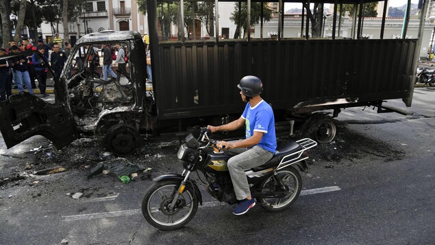 Une carcasse de camion brûlé à côté d'un homme sur une motocyclette.