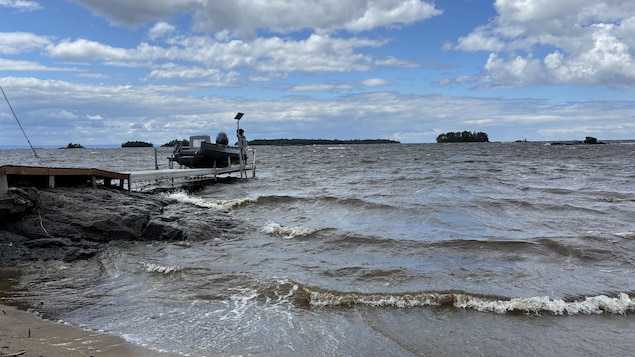 Une étendue d'eau agitée par des vagues borde un rivage rocheux et sablonneux. Sur la gauche, un quai soutient un bateau à moteur hors-bord. À l'horizon, des îles boisées se dessinent sous un ciel parsemé de nombreux nuages blancs.