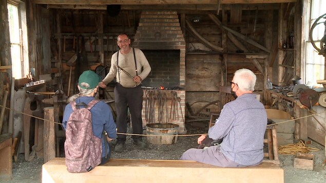 Une Forge du village historique acadien de la Nouvelle-Écosse : deux visiteurs sont en face d'un homme debout habillé avec le style vestimentaire des années 1900.