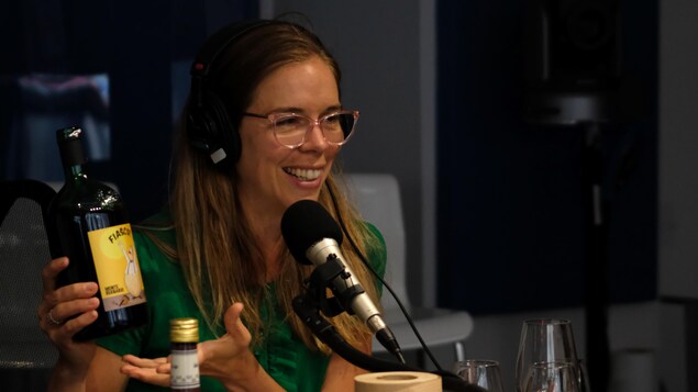 Une femme avec des lunettes et une chemise verte tient une bouteille de vin en parlant à un micro dans un studio de radio.