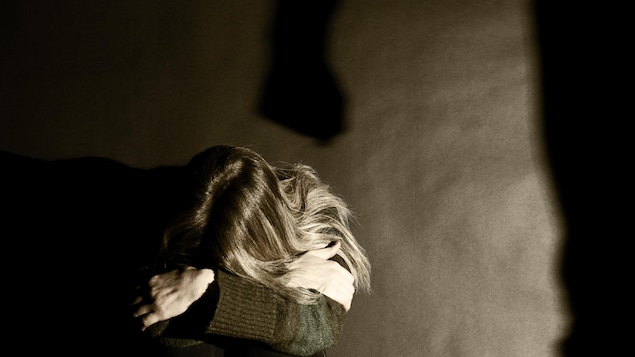 Une femme assise la tête entre les bras et l'ombre d'un poing fermé sur le mur au-dessus d'elle.