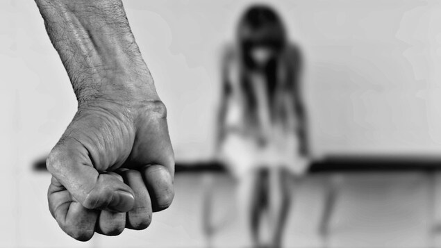 Un poing serré au premier plan et une femme assise, tête baissée, en arrière-plan.