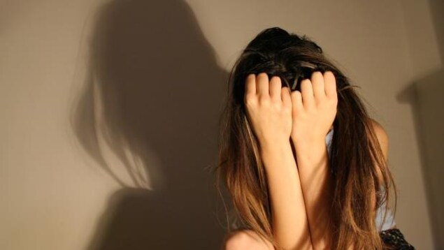 Une femme se couvre le visage dans un coin d'une pièce avec son ombre au dessus d'elle.