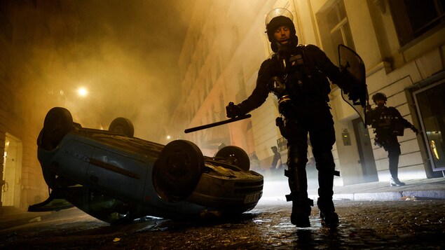 Un policier avec son équipement anti-émeute devant une voiture renversée.