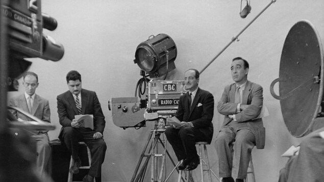 Sur des tabourets, des journalistes, dont Raymond Charette et Lucien Côté, révisent leurs notes entourés de caméras et d'autre équipement technique.
