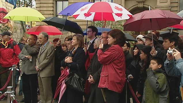 Des personnes qui attendent sous la pluie derrière un ruban rouge. La plupart ont des parapluies et certains tiennent un appareil photo. Des personnes qui attendent sous la pluie derrière un ruban rouge. La plupart ont des parapluies et certains tiennent un appareil photo.