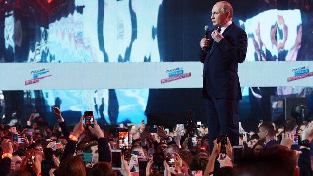 Vladimir Poutine se tient debout sur une scène, micro à la main. Devant lui, un parterre rempli de jeunes qui tiennent leur téléphone portable bien haut pour pouvoir le prendre en photo.