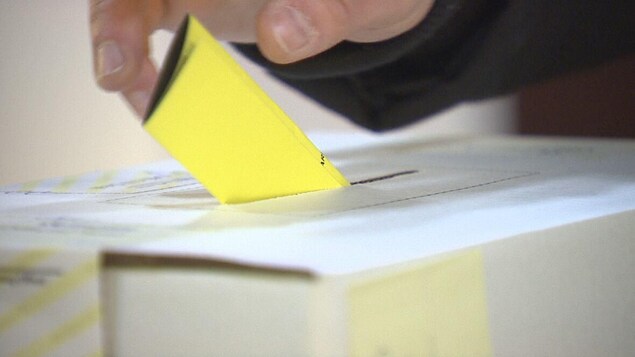Une main dépose dans la fente d'une boîte de scrutin un bulletin de vote jaune plié en trois parties.