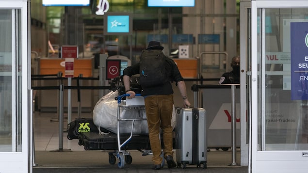 Un voyageur entre dans l'aéroport, chargé de bagages, en poussant un chariot.
