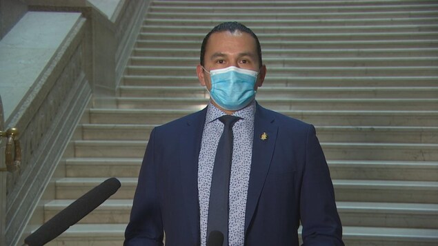 Wab Kinew porte un masque bleu, une chemise blanche à pois bleus, une cravate bleue et un veston bleu. Il est devant des escaliers au palais législatif du Manitoba.