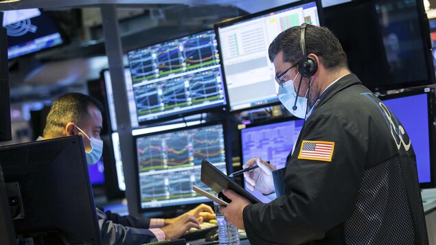 Deux hommes à la Bourse de New York devant des écrans sur lesquels s'affichent des graphiques.
