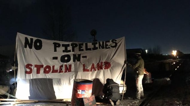 Une voie ferrée bloquée par des voitures. Des personnes installent une banderole où il est écrit: pas de pipeline sur une terre volée.