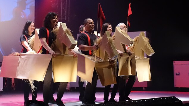 Un groupe de comédiens sont debout souriants sur scène, déguisés en cheval de carton, le costume tenant à l'aide de bretelles rouges.