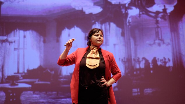 La comédienne Dominique Pétin, habillée d'un pantalon et d'un t-shirt noir, avec un veston rouge. Sa main droite posée sur la hanche, elle tient un cigare dans sa main gauche. En arrière-plan, une projection d'un grand salon de style victorien. La comédienne Dominique Pétin, habillée d'un pantalon et d'un t-shirt noir, avec un veston rouge. Sa main droite posée sur la hanche, elle tient un cigare dans sa main gauche. En arrière-plan, une projection d'un grand salon de style victorien.