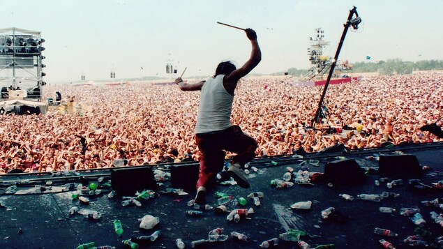 Un homme danse avec des baguettes de batterie sur une scène jonchée de bouteilles d'eau, devant une foule monstre à l'extérieur. 