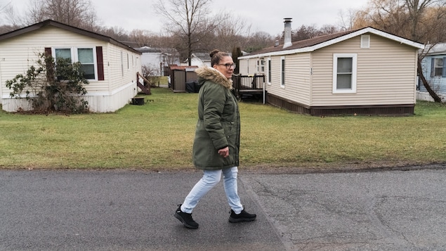 Yvonne marche sur une rue asphaltée et porte un manteau d'hiver avec un col de fourrure. À sa gauche, on voit deux maisons mobiles de couleur beige.