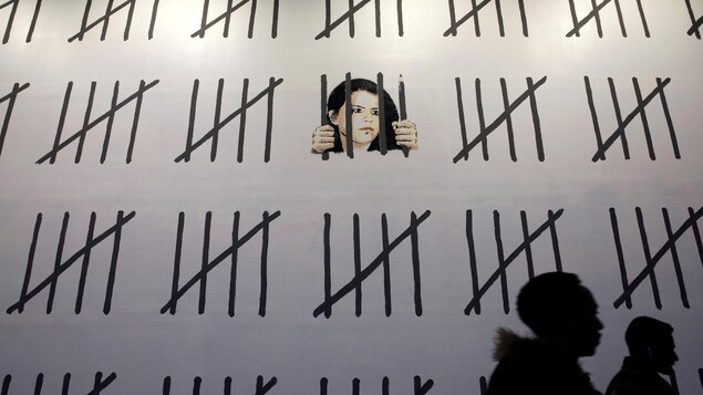 Deux passants marchent devant un mur blanc. Le visage de la journaliste et artiste Zehra Dogan y a été dessiné par Banksy derrière des barreaux, dont un est aussi un crayon. D'autres traits noirs représentent les jours passés en prison par la Turque. Deux passants marchent devant un mur blanc. Le visage de la journaliste et artiste Zehra Dogan y a été dessiné par Banksy derrière des barreaux, dont un est aussi un crayon. D'autres traits noirs représentent les jours passés en prison par la Turque.