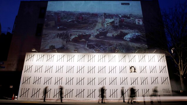 Des passants flous marchent devant la murale géante de Banksy, à New York. Sur ce mur blanc, des traits noirs représentent les jours passés en prison par la journaliste. On voit aussi son visage derrière quatre traits qui prennent l'aspect d'une prison. Des passants flous marchent devant la murale géante de Banksy, à New York. Sur ce mur blanc, des traits noirs représentent les jours passés en prison par la journaliste. On voit aussi son visage derrière quatre traits qui prennent l'aspect d'une prison.