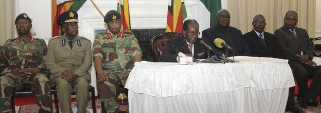 Robert Mugabe est assis derrière un bureau sur lequel se trouvent des micros. Des hommes en uniforme sont assis à côté de lui, à gauche. D'autres en complet sont assis à droite. Robert Mugabe est assis derrière un bureau sur lequel se trouvent des micros. Des hommes en uniforme sont assis à côté de lui, à gauche. D'autres en complet sont assis à droite.