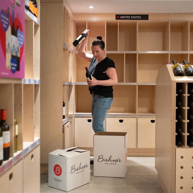 Dans un magasin d'alcool, devant des étagères en bois, une femme tient une bouteille de vin dans chaque main. Les tablettes sous l'écriteau « États-Unis » sont vides et des boîtes de carton sont sur le sol.