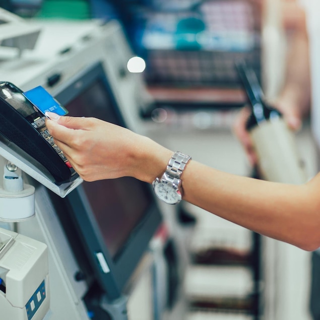 Une personne d&eacute;pose une carte de paiement sur le dessus terminal de paiement &agrave; une caisse libre-service dans un commerce.