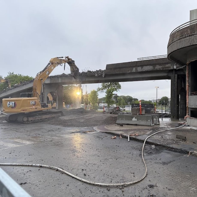 Une pelle mécanique est en train de démolir une structure de béton qui surplombe une autoroute. 