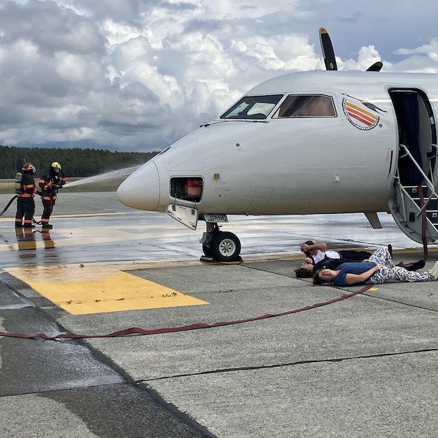 Des pompiers arrosent un avion. Des figurants sont couchés au sol.