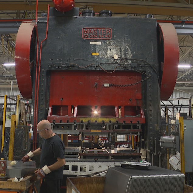Vue intérieure d'une usine montrant un homme chauve portant des lunettes et des gants, se penchant sur une table de travail. En arrière-plan domine une presse mécanique d'estampage noire et rouge de marque "MINSTER". L'environnement est celui d'un atelier industriel avec des outils et des structures métalliques.