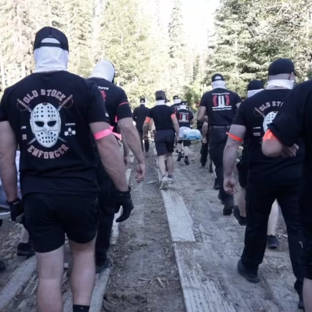 Des hommes cagoulés portant des t-shirts noirs et qui transportent des lits de camp sur un pont en bois dans une forêt.